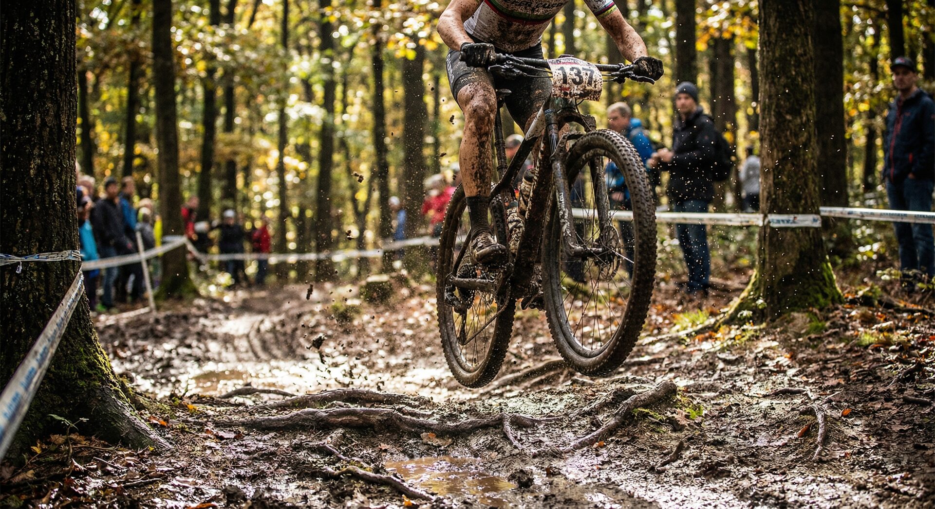 Mountain bike rider racing through muddy forest trail — bib numbers obscured by mud and dynamic positioning
